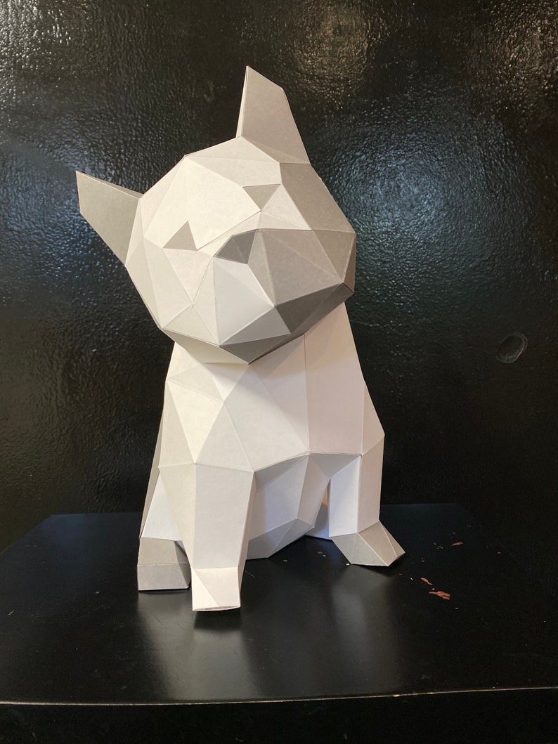 May include: A white and grey papercraft model of a French bulldog sitting on a black surface. The model is made of folded paper and has a geometric design.