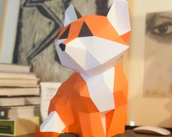 Pode incluir: Uma escultura de papel de uma raposa de cor laranja, branca e preta. A raposa está sentada com a cabeça inclinada para o lado e os olhos fechados.