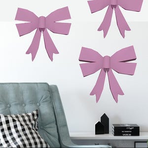 May include: Three large pink papercraft bows on a white wall. The bows are in the shape of a traditional bow with a center knot and two loops on each side. The bows are hanging on a wall with a light blue armchair and a white shelf with books and decorative objects.