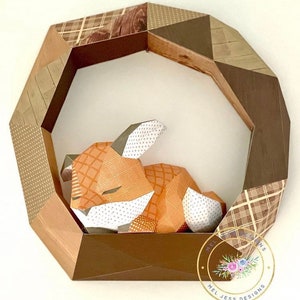 K&ouml;nnte beinhalten: Ein brauner geometrischer Rahmen aus Holz mit einem gemusterten Papierdesign. Ein Papier-Fuchs befindet sich im Rahmen und liegt mit dem Kopf auf den Pfoten.
