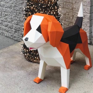 Op de afbeelding: Een geometrische papieren beagle hond sculptuur. De hond heeft oranje, witte en zwarte driehoekige panelen. De oren en de kop zijn oranje, met witte en zwarte accenten op het gezicht. De hond staat op een betonnen oppervlak.