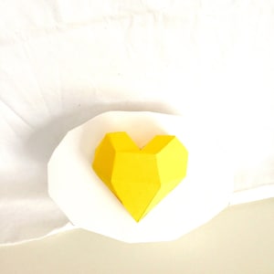 May include: A bright yellow, geometric heart shape, resting on a white, petal-like form. The heart is constructed from folded paper, showcasing a faceted design. The background is a soft, off-white fabric.