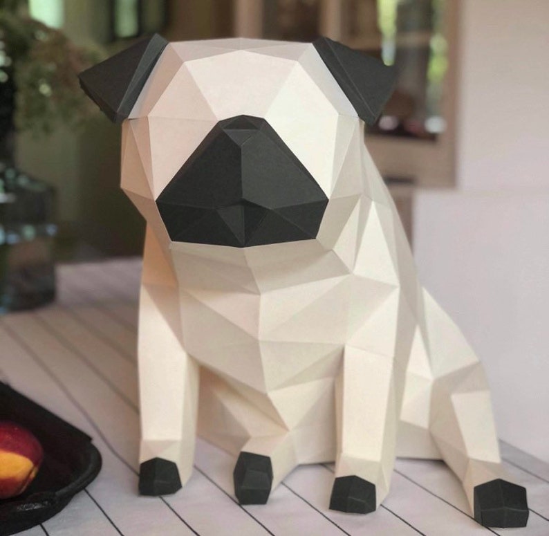Op de afbeelding: Een wit papieren pug-beeldje met zwarte oren, neus en poten. Het beeldje zit en heeft zijn hoofd lichtjes naar de zijkant gekanteld.