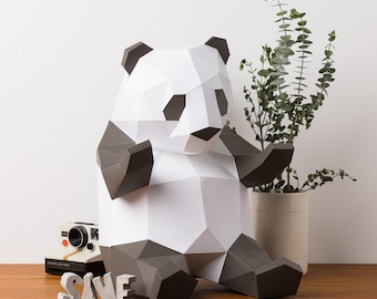 May include: A white and gray papercraft panda sculpture sitting on a wooden surface. The panda is sitting with its arms outstretched and has a black nose and eyes.
