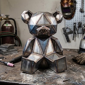 Puede incluir: Escultura geométrica de metal de un oso, construida con acero soldado. El oso tiene un diseño facetado con líneas de soldadura visibles, y está sobre una superficie metálica. La escultura está en un taller.