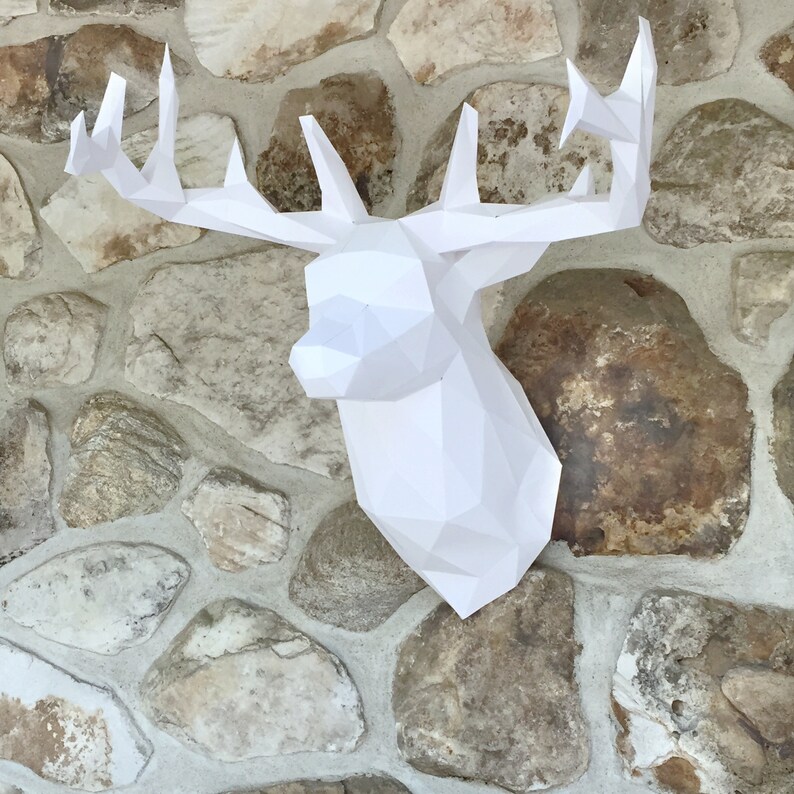 May include: White paper deer head sculpture mounted on a stone wall. The deer head is made of folded paper and has a geometric design.