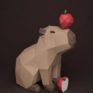 May include: A low-poly papercraft capybara sculpture in shades of tan and brown. The figure has a geometric, faceted design. A small, red, faceted paper apple rests on its head, and a halved red paper apple sits on the ground by its front feet.