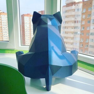 May include: A blue papercraft sculpture of a sitting cat. The sculpture is made of folded paper and has a geometric design. The cat is sitting on a white surface with a green surface in the background.