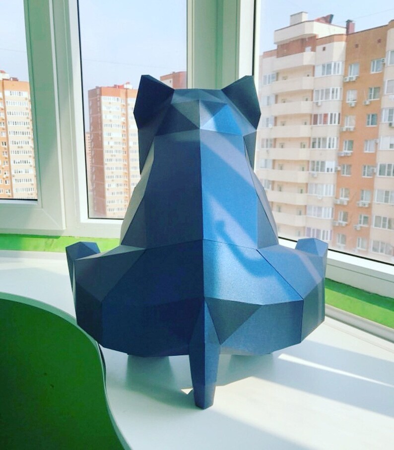 May include: A blue papercraft sculpture of a sitting cat. The sculpture is made of folded paper and has a geometric design. The cat is sitting on a white surface with a green surface in the background.
