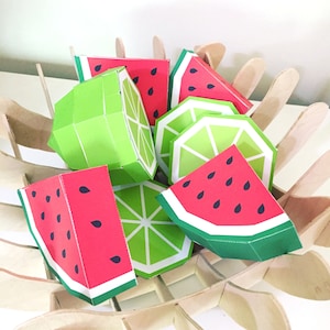 Puede incluir: Una colección de cajas de papel con forma de fruta en una cesta de madera. Las cajas tienen forma de limas y sandías y están decoradas con colores verde y rojo.