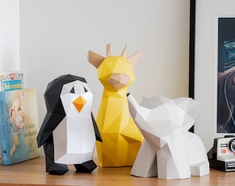 May include: Three geometric paper animal sculptures: a black and white penguin, a yellow giraffe, and a gray elephant. The figures are displayed on a wooden surface, with a book and a framed picture in the background.