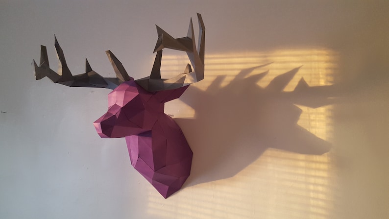 May include: A purple papercraft deer head with large antlers mounted on a white wall. The deer's shadow is cast on the wall behind it.