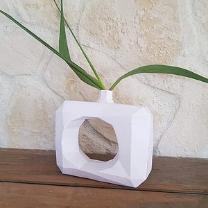 Puede incluir: Jarrón de cerámica blanco geométrico con una gran abertura circular, con un diseño sencillo. El jarrón está lleno de tallos verdes con hojas.