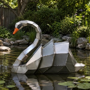 May include: A geometric metal swan sculpture constructed from multiple silver-toned steel panels with visible welded seams. The swan features a long, curved neck and a bright orange beak, floating on the surface of a dark garden pond.