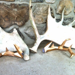 May include: Two gold-colored, geometric deer antlers with purple accents, resting on a white antler against a stone wall background.