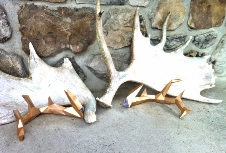 May include: Two gold-colored, geometric deer antlers with purple accents, resting on a white antler against a stone wall background.