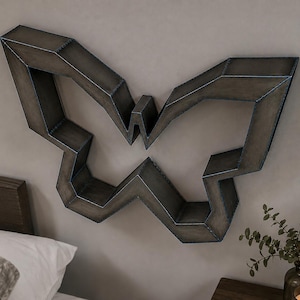 Puede incluir: Un estante de pared con forma de mariposa construido en metal gris oscuro con costuras soldadas visibles a lo largo de los bordes. El estante presenta un diseño geométrico y angular con múltiples superficies planas para exhibición.