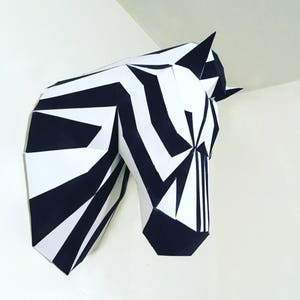 May include: A geometric papercraft zebra head sculpture, featuring bold black and white stripes. The artwork is mounted on a white wall, showcasing the animal's head and part of its neck. The design is modern and angular.