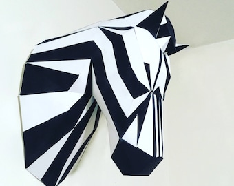 May include: A geometric papercraft zebra head sculpture, featuring bold black and white stripes. The artwork is mounted on a white wall, showcasing the animal's head and part of its neck. The design is modern and angular.