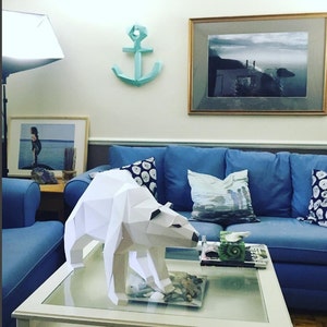May include: A white papercraft polar bear sculpture on a glass-top coffee table in a living room. The bear is standing on a bed of small white stones. The room has a blue couch with pillows, a framed photo on the wall, and a blue paper anchor hanging on the wall.