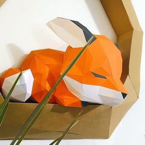 K&ouml;nnte beinhalten: Eine Papierkunst-Fuchs-Skulptur in Orange, Wei&szlig; und Schwarz, die auf einem braunen Kartonhintergrund schl&auml;ft.