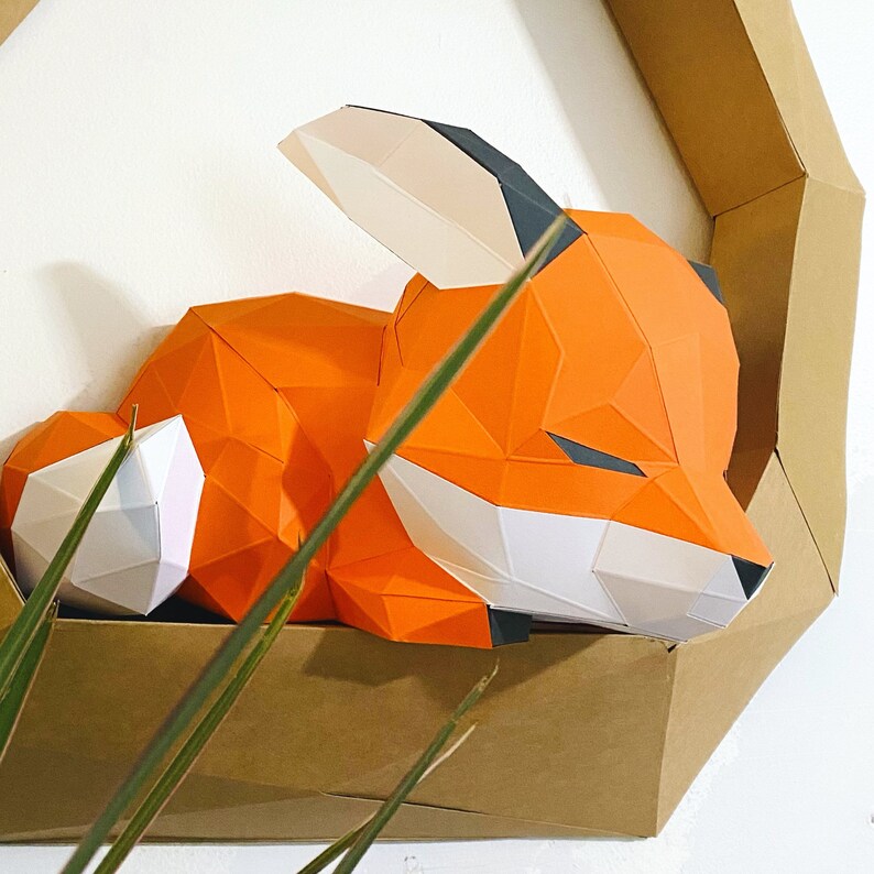 K&ouml;nnte beinhalten: Eine Papierkunst-Fuchs-Skulptur in Orange, Wei&szlig; und Schwarz, die auf einem braunen Kartonhintergrund schl&auml;ft.