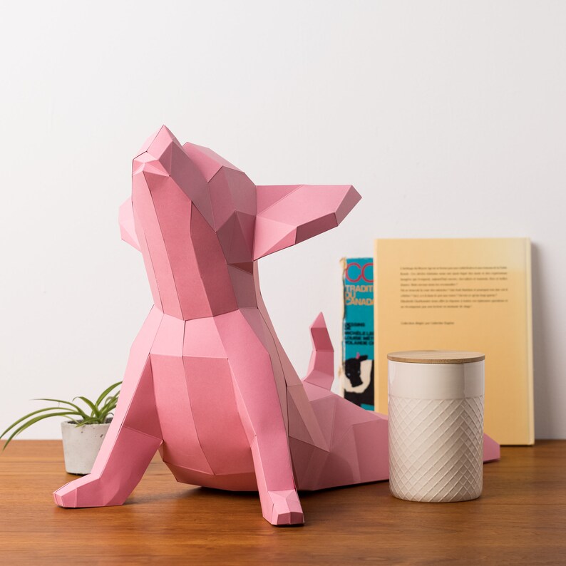 May include: A pink origami paper chihuahua sculpture in a seated position with its head tilted back. The sculpture is on a wooden table with a white ceramic jar and a book in the background.