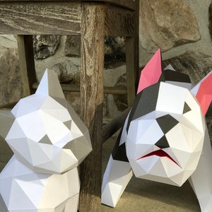 May include: Two papercraft dog sculptures, one white and one black and white, are posed next to a wooden chair. The white dog is sitting, while the black and white dog is standing with its head tilted down.