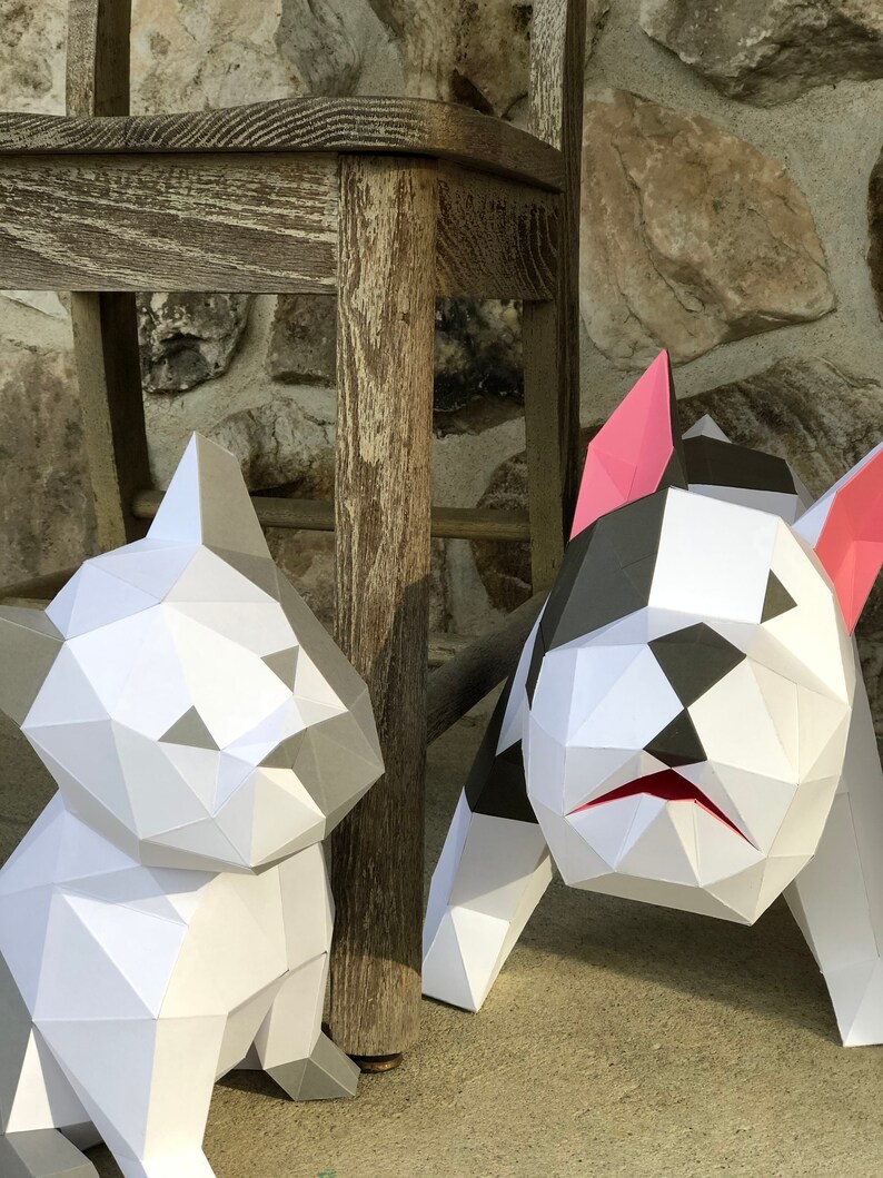 May include: Two papercraft dog sculptures, one white and one black and white, are posed next to a wooden chair. The white dog is sitting, while the black and white dog is standing with its head tilted down.