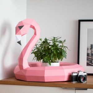 Pode incluir: Uma escultura de flamingo rosa de papel com um vaso branco dentro. O flamingo tem a forma de um anel e um pescoço comprido. Uma câmera vintage está sentada em uma superfície de madeira na frente do flamingo.