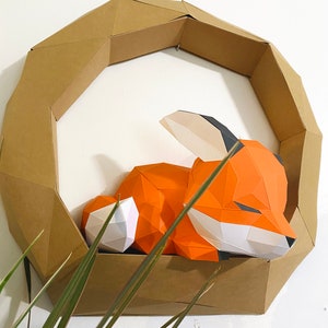 K&ouml;nnte beinhalten: Eine Papierkunst-Fuchs-Skulptur in Orange, Wei&szlig; und Schwarz, die in einem braunen geometrischen Kartonrahmen schl&auml;ft.