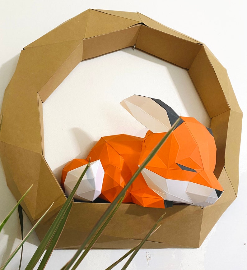 K&ouml;nnte beinhalten: Eine Papierkunst-Fuchs-Skulptur in Orange, Wei&szlig; und Schwarz, die in einem braunen geometrischen Kartonrahmen schl&auml;ft.
