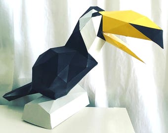 May include: A papercraft toucan sculpture with a black body, white head, and yellow beak. The toucan is standing on a white base.