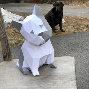 May include: A white and grey papercraft model of a dog sitting on a wooden chair. The dog is made of folded paper and has a realistic appearance.