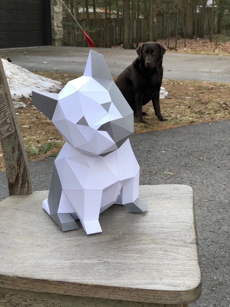 May include: A white and grey papercraft model of a dog sitting on a wooden chair. The dog is made of folded paper and has a realistic appearance.