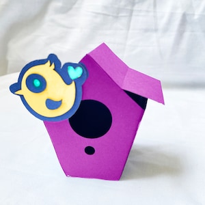 May include: A purple paper birdhouse with a yellow bird cutout on the front. The bird has a blue eye and a small blue heart.