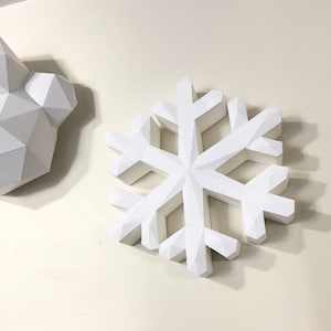 May include: A large, white, geometric snowflake decoration with a faceted design. The snowflake is mounted on a light-colored wall, casting a shadow. A partial view of a white geometric bear head is visible to the left.