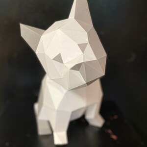 May include: A white papercraft model of a French bulldog sitting on a black surface. The model is made of folded paper and has a geometric design.