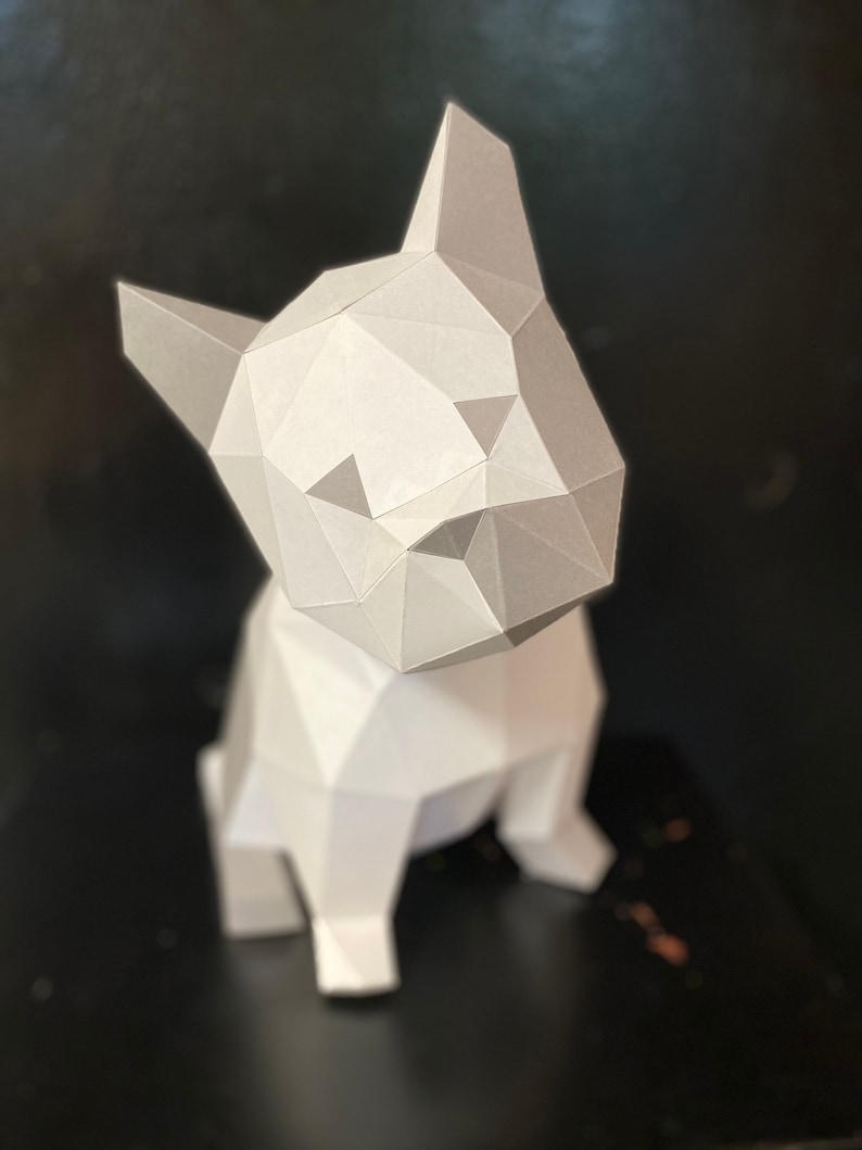 May include: A white papercraft model of a French bulldog sitting on a black surface. The model is made of folded paper and has a geometric design.