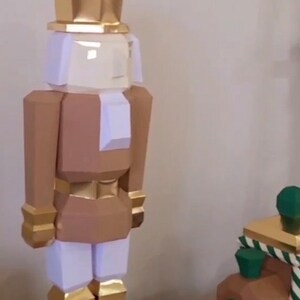 Puede incluir: Una figura de cascanueces de papel artesanal de color dorado y marrón con una cara blanca y un sombrero dorado. El cascanueces está de pie sobre una base de madera. Un tren de papel artesanal verde y marrón está en el fondo.