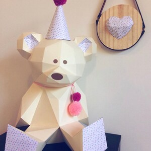May include: A geometric paper bear sculpture with a party hat and pink pom-poms. The bear is cream-colored with a geometric design. The hat is white with a pattern of small gray hearts. A wooden wall hanging with a paper heart is also visible.