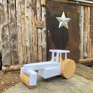 May include: A white cardboard model of a steamroller with brown cardboard wheels. The model is sitting on a large rock in front of a wooden building with a sign that reads "Welcome to the Cabin".