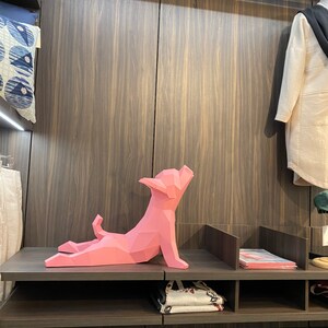 May include: A pink origami-style dog sculpture on a shelf in a closet. The dog is in a yoga pose with its legs stretched out and its head tilted back.
