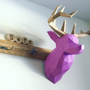 May include: A pink and gold papercraft deer head mounted on a wall. The deer head is made of folded paper and has a geometric design. The deer head is mounted on a wooden ruler with the word "Sofas" written on it.