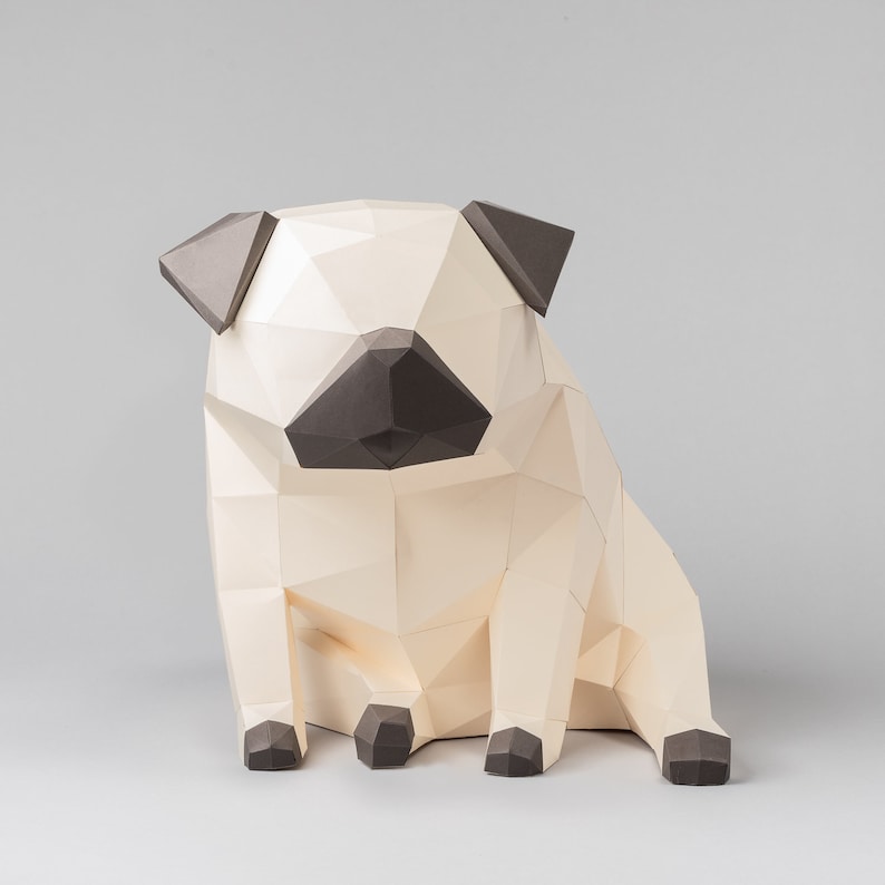Op de afbeelding: Een beige papieren pug-hondensculptuur met bruine oren en een bruine neus. Het beeldje staat op een grijze ondergrond.