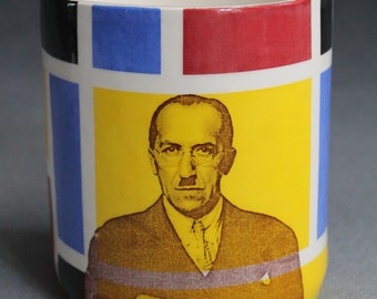 May include: A white ceramic mug with a geometric design featuring red, blue, black, and yellow squares. The mug has a portrait of a man with glasses and a mustache in the center.
