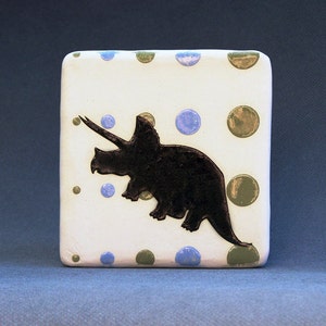 May include: A white ceramic tile with a black silhouette of a triceratops dinosaur. The tile is decorated with blue and green polka dots.