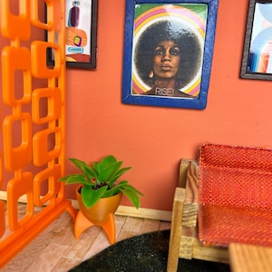 May include: A miniature mid-century modern living room scene. The orange walls are adorned with framed art, including a portrait with the text "SUPER SOUL RISE!". A small orange planter holds a green plant. An orange room divider and a red couch complete the scene.