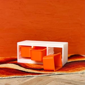 May include: A white storage unit with three compartments, two containing bright orange bins. One orange bin is pulled out. The unit sits on a rug with orange, brown, and cream patterns, against an orange wall.
