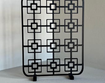 May include: A black decorative screen with a geometric square pattern. The screen is rectangular with rounded corners and stands on two small feet. The design features a repeating pattern of squares within squares, creating an open, airy aesthetic.